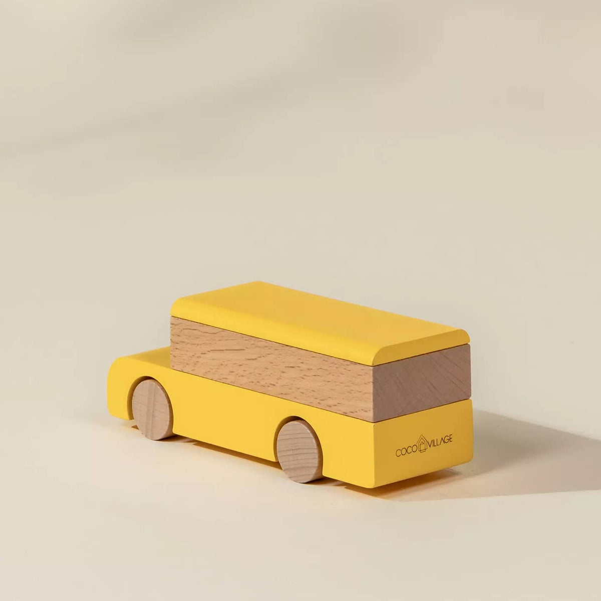 A wooden toy bus by Cocoon Block, featuring a bright yellow painted body and natural wood wheels and side panel, set against a warm beige background.