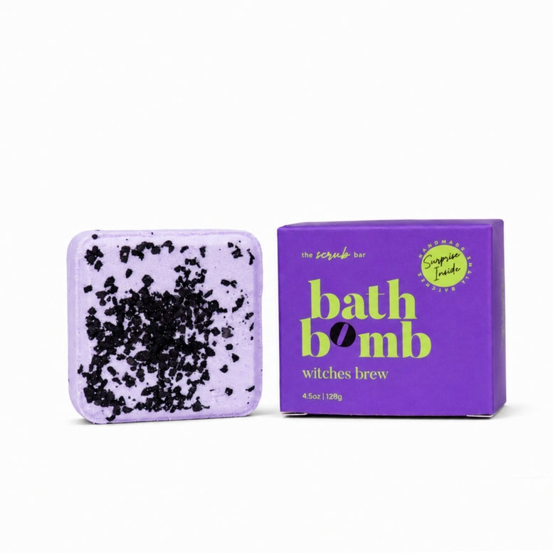A lavender bath bomb with black speckles shown next to its purple packaging box on a white background. The box is branded 'the scrubs by bath bomb' and labeled 'witches brew' with 'sugar cane' in a green circle.