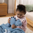 A young child sitting on a floor in a bedroom, holding a light blue plush dinosaur toy under a blue blanket.