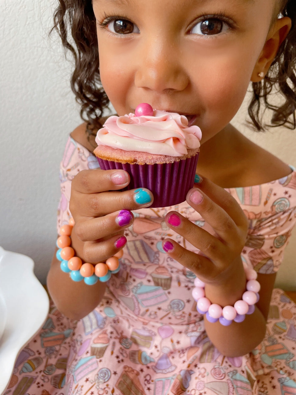 Child holding a pink cupcake with colorful frosting, wearing multiple bracelets.