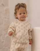 A toddler with curly light brown hair wearing a cream-colored waffle-knit long-sleeve top and matching pants with a small floral pattern, standing indoors next to a white chair.