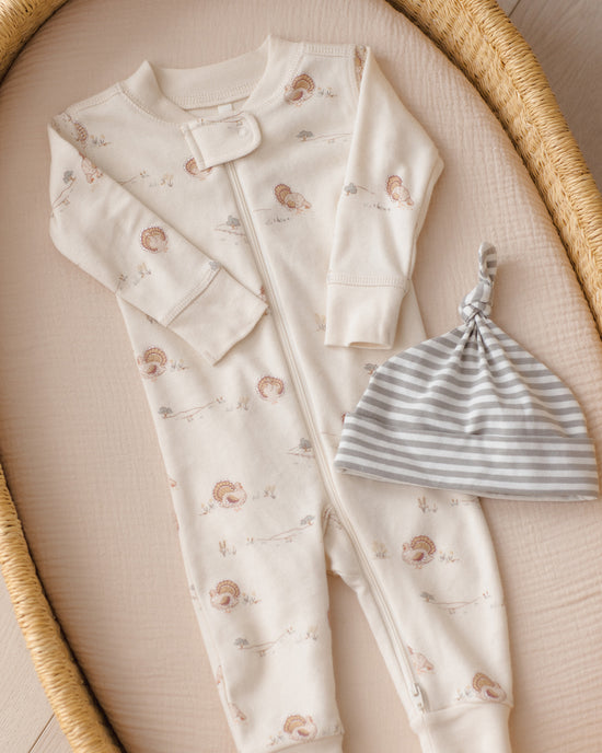 A baby one-piece footless sleeper in cream with a small hedgehog print, laid flat on a beige cushion inside a woven tray, next to a gray and white striped knit beanie.