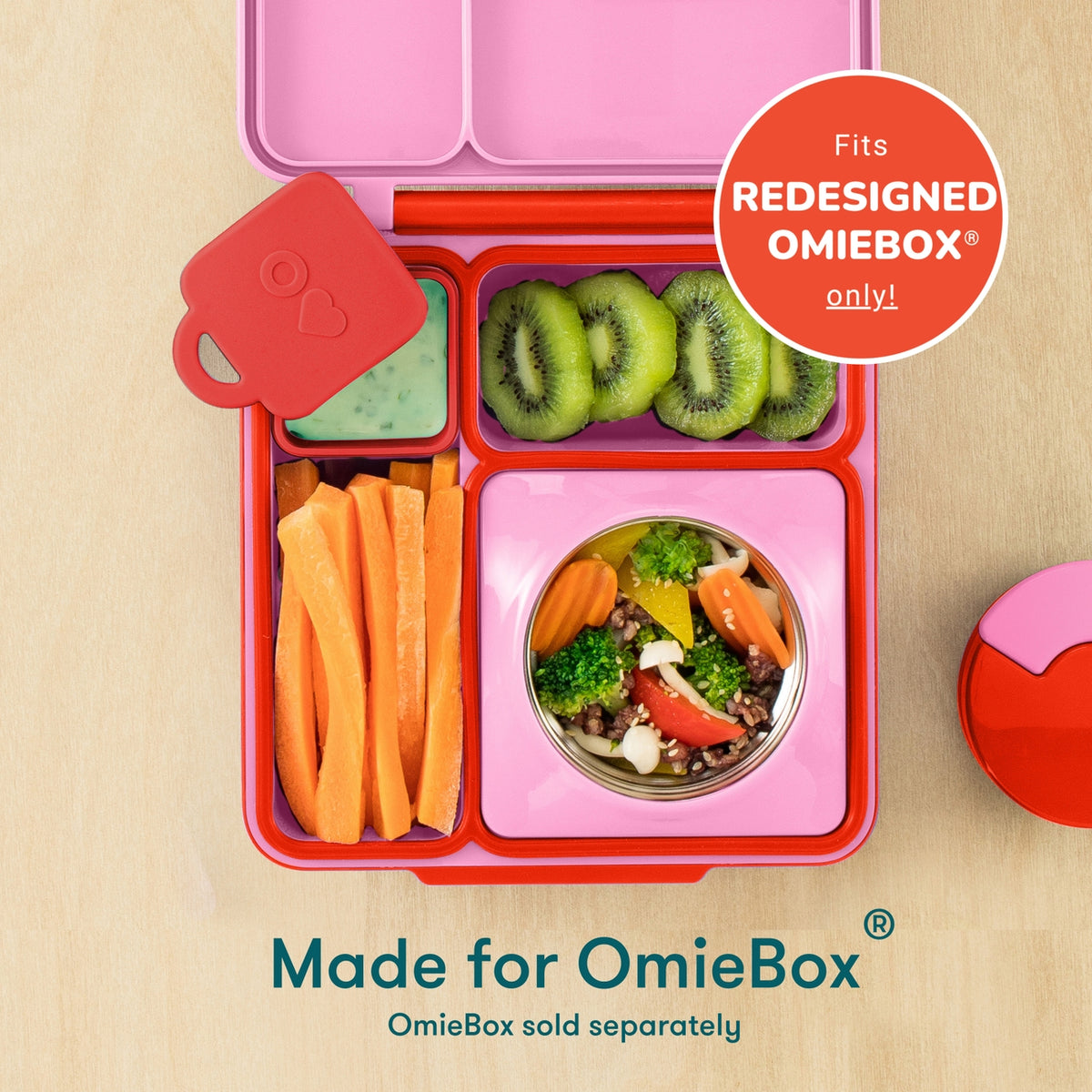 A pink and red OmieBox lunch box filled with sliced kiwi, carrot sticks, a green dip, and a small salad bowl, shown on a light wood background with text noting it fits the redesigned OmieBox only.