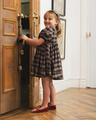 A young girl with blonde hair stands in a doorway wearing a brown and black plaid dress and red shoes, holding the door open with one hand.