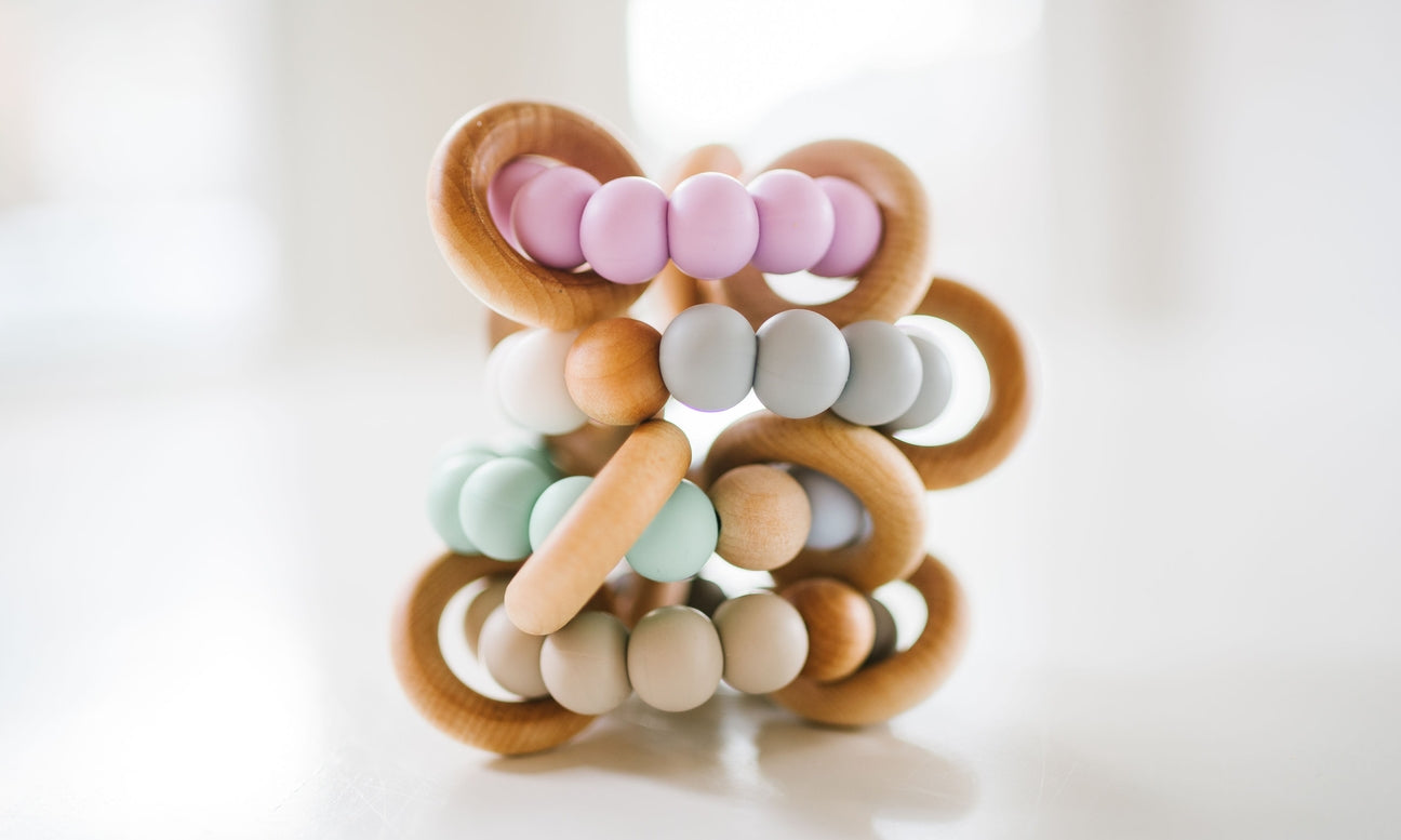 A wooden bead teether toy with interlocking natural wood rings and pastel-colored beads in pink, mint green, light blue, beige, and white, photographed on a soft white background.