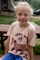 A young girl with blonde hair blowing in the wind is wearing a light pink graphic t-shirt with a brown cat design and the text 'BRING ME All', paired with blue pants, sitting outdoors on a wooden bench.