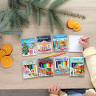 A top-down view of a child's hands using a pink pencil on a white spiral notebook, set on a light wooden table alongside six colorful children's activity books, pine branches, and orange slices.