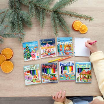 A top-down view of a child's hands using a pink pencil on a white spiral notebook, set on a light wooden table alongside six colorful children's activity books, pine branches, and orange slices.