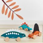 Three wooden dinosaur toys on a light surface with a decorative leaf in the background.