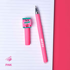 A pink retractable pen and matching pink highlighter with a cartoon character charm labeled 'STROKES' are arranged on a lined notebook page next to a solid pink background. The word 'PINK' appears in the bottom-left corner.