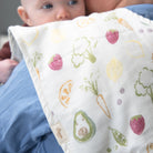 Baby wrapped in a blanket with fruit and vegetable patterns, held by an adult.