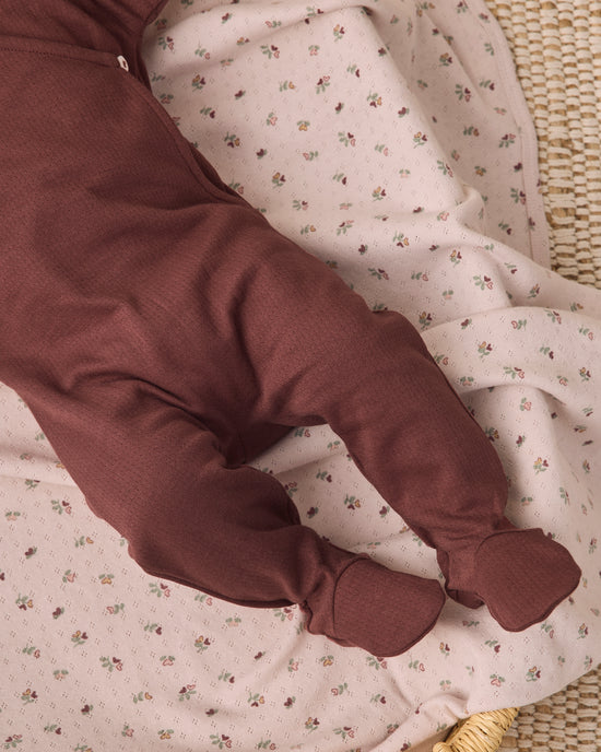 A close-up of a baby's legs wearing a dark brown footed sleeper, lying on a light pink muslin blanket with a small floral print.
