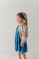 A young girl wearing a tiered blue dress, a beige cardigan with colorful tulip patterns, and a blue and white gingham headband, standing in profile against a plain white studio background.