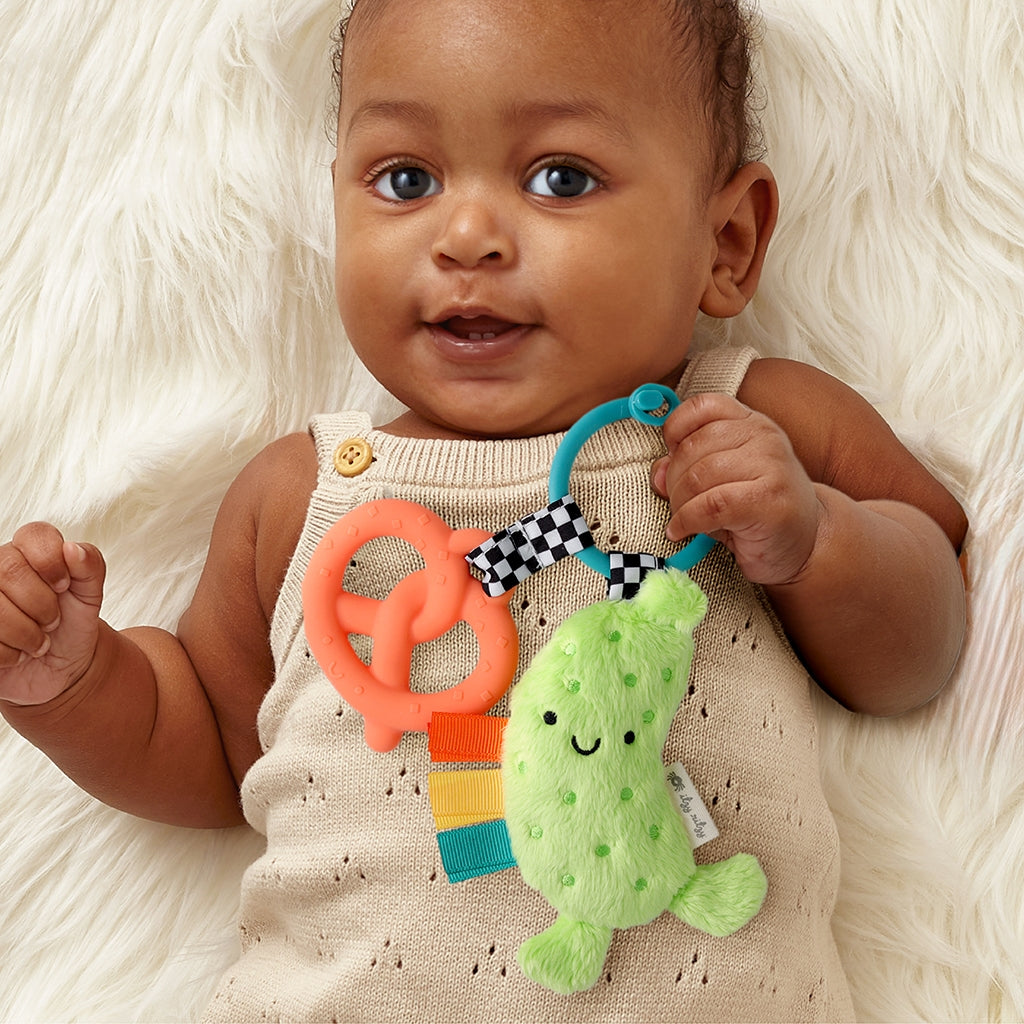 Baby holding a colorful teething toy with a green fish attachment on a white fluffy surface