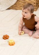 A baby wearing a brown ribbed sleeveless top sits on a cream textured blanket, holding a small yellow toy while nearby are two flower-shaped stacking toys in orange and yellow.