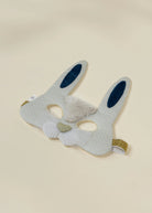 A white fabric bunny mask with dotted pattern, featuring blue inner ears, a fluffy white faux fur nose, and gold glitter trim on the ear tips and side straps, shown on a light beige background.
