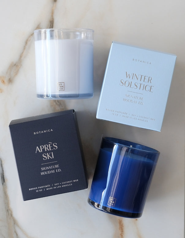 Two Botanica candles and their packaging boxes arranged on a light marble surface. One candle is in a clear glass jar with white wax and a blue tint near the bottom, next to a pale blue box labeled Winter Solstice.