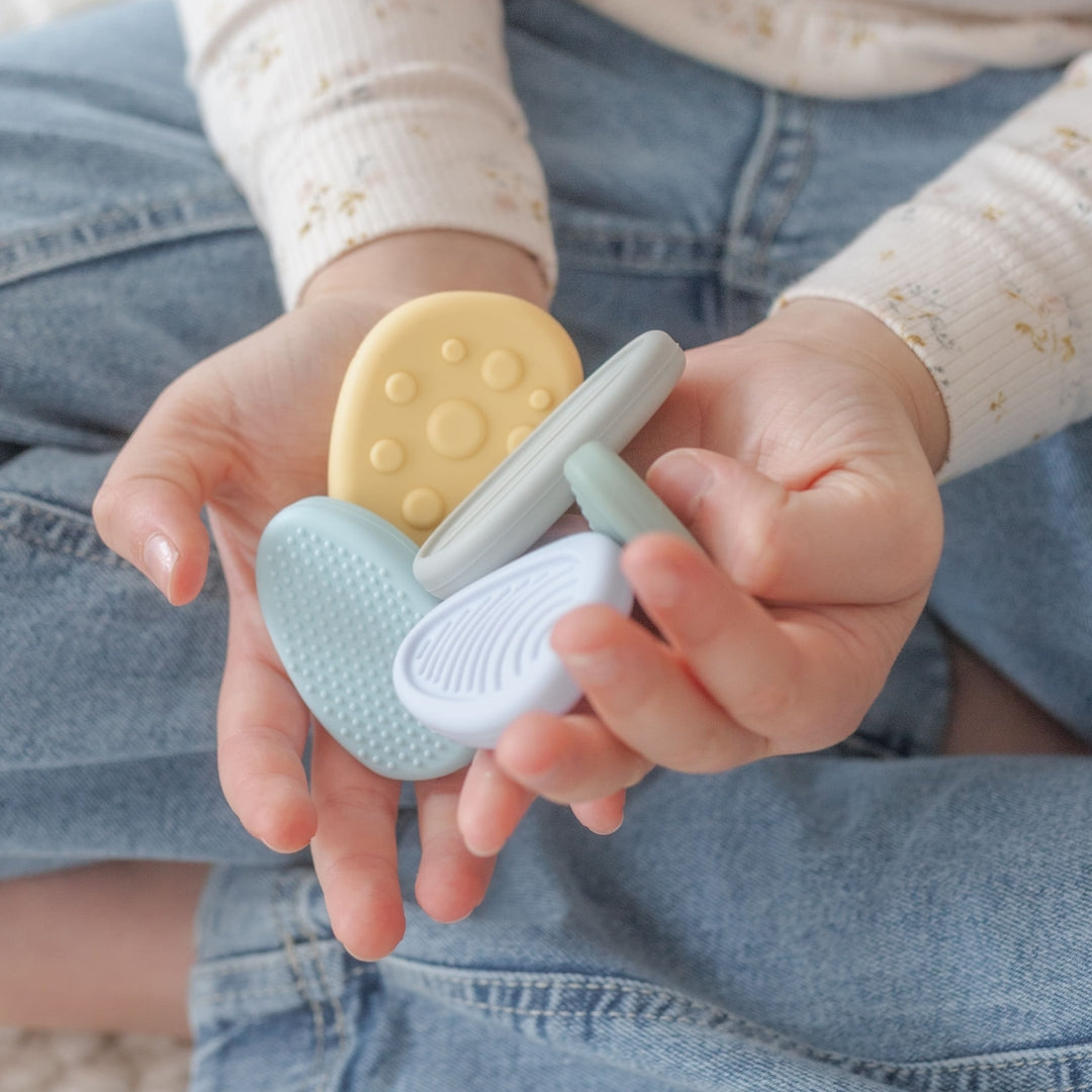 Baby hands holding two silicone teething rings, one yellow and one light blue, against a denim background.