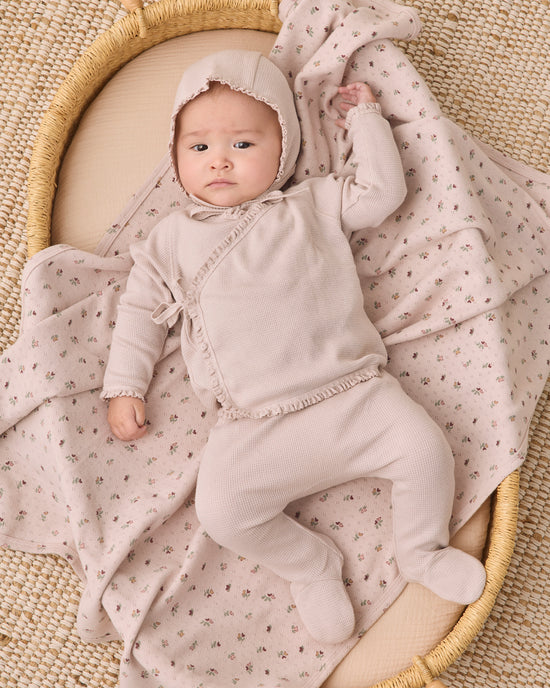 A baby wearing a light beige footed onesie and bonnet lies on a cream woven mat, wrapped in a matching beige and pink floral blanket.