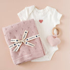Baby onesie with heart design, pink knitted blanket, and pink teething ring on a beige background