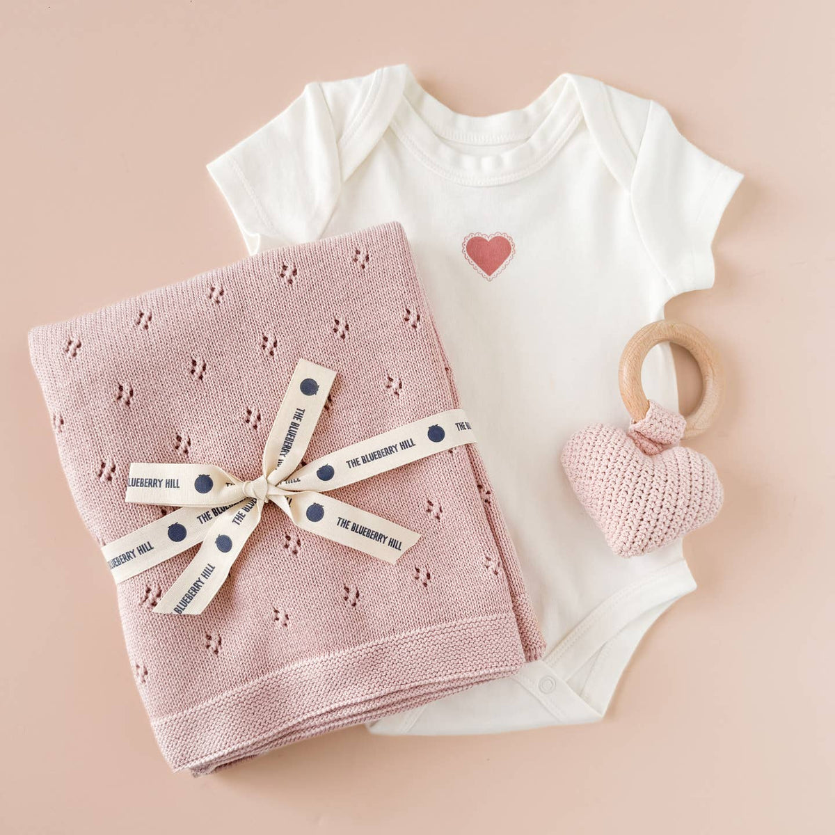 Baby onesie with heart design, pink knitted blanket, and pink teething ring on a beige background