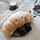 Plush toy shaped like a bread roll on a chair with a cup of coffee and books in the background.