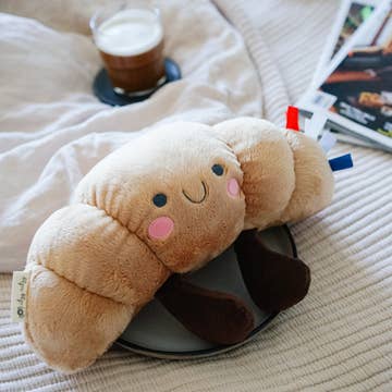 Plush toy shaped like a bread roll on a chair with a cup of coffee and books in the background.