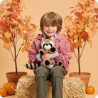 A young child in a pink hoodie holding a plush Raccoon by Warmies, sitting on a bale of hay with pumpkins and autumn leaves in the background.