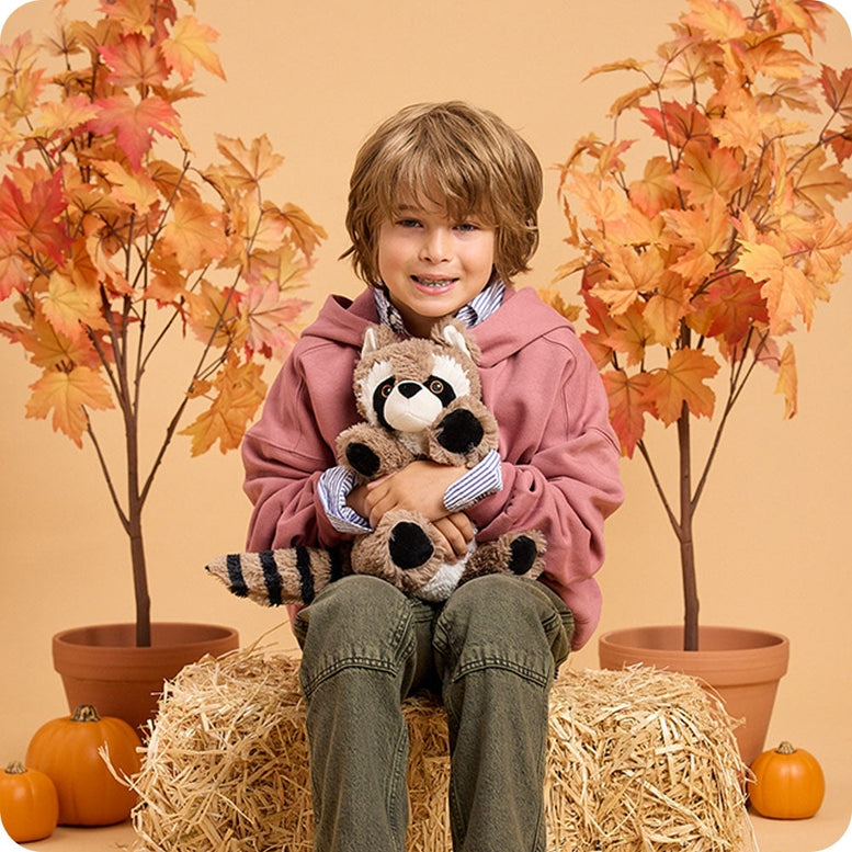 A young child in a pink hoodie holding a plush Raccoon by Warmies, sitting on a bale of hay with pumpkins and autumn leaves in the background.