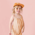 A young child smiling and wearing a peach-orange gingham sleeveless romper and a matching orange cap, posed against a solid dusty pink background.