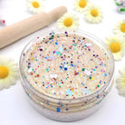 Jar of beige-colored slime with colorful sprinkles on a white surface with daisy flowers and a rolling pin.