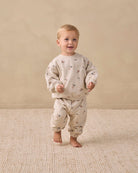 A toddler standing barefoot on a textured beige rug against a warm beige wall, wearing a cream-colored long-sleeve top and matching pants with a small scattered animal print.