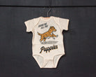 An off-white short-sleeve baby bodysuit laid flat on a textured surface, featuring a graphic of a yellow and white puppy with the text 'BRING ME ALL THE Puppies'.