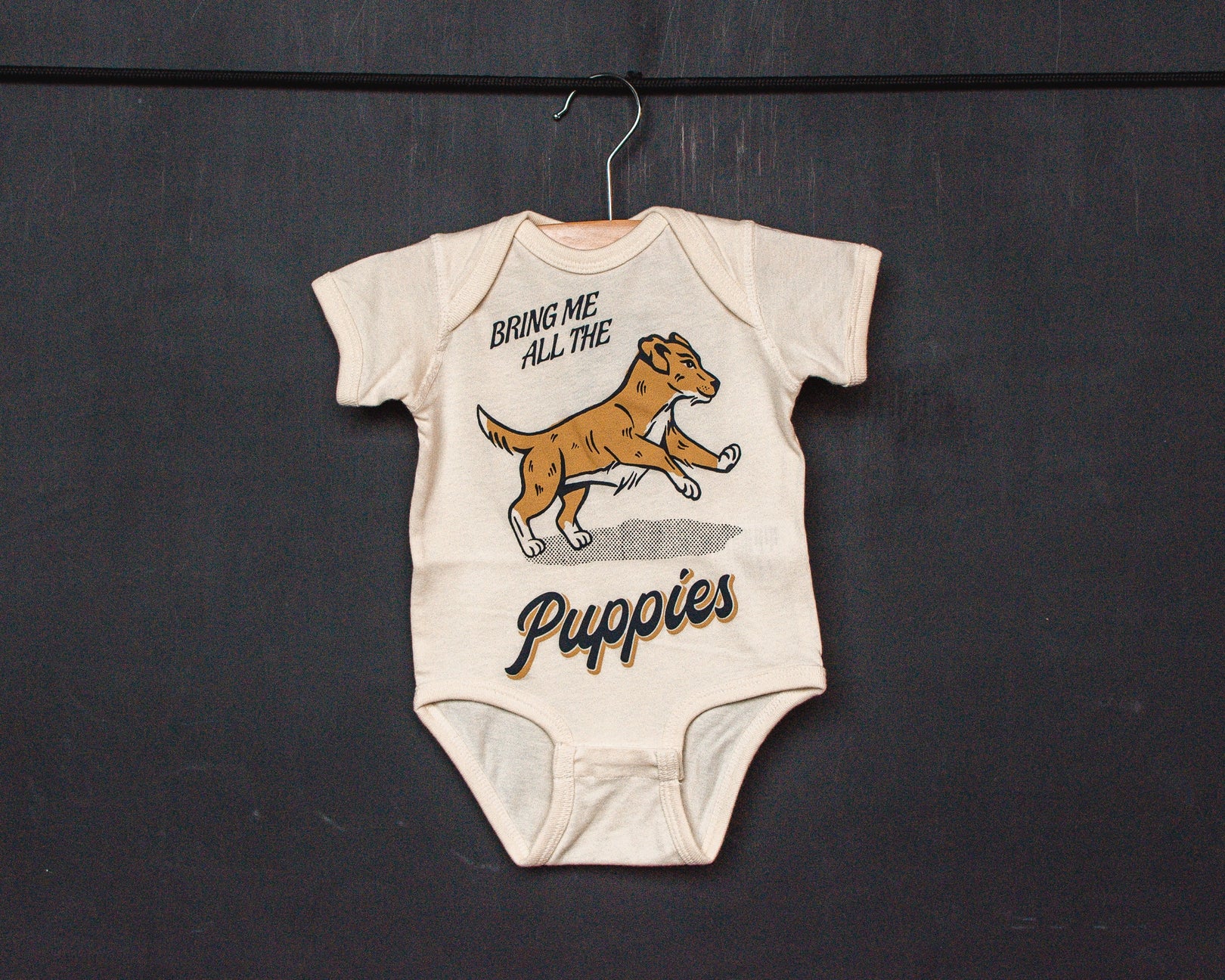 An off-white short-sleeve baby bodysuit laid flat on a textured surface, featuring a graphic of a yellow and white puppy with the text 'BRING ME ALL THE Puppies'.