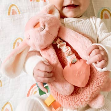 Baby holding a pink plush toy with a heart-shaped rattle on a white blanket with rainbow patterns.
