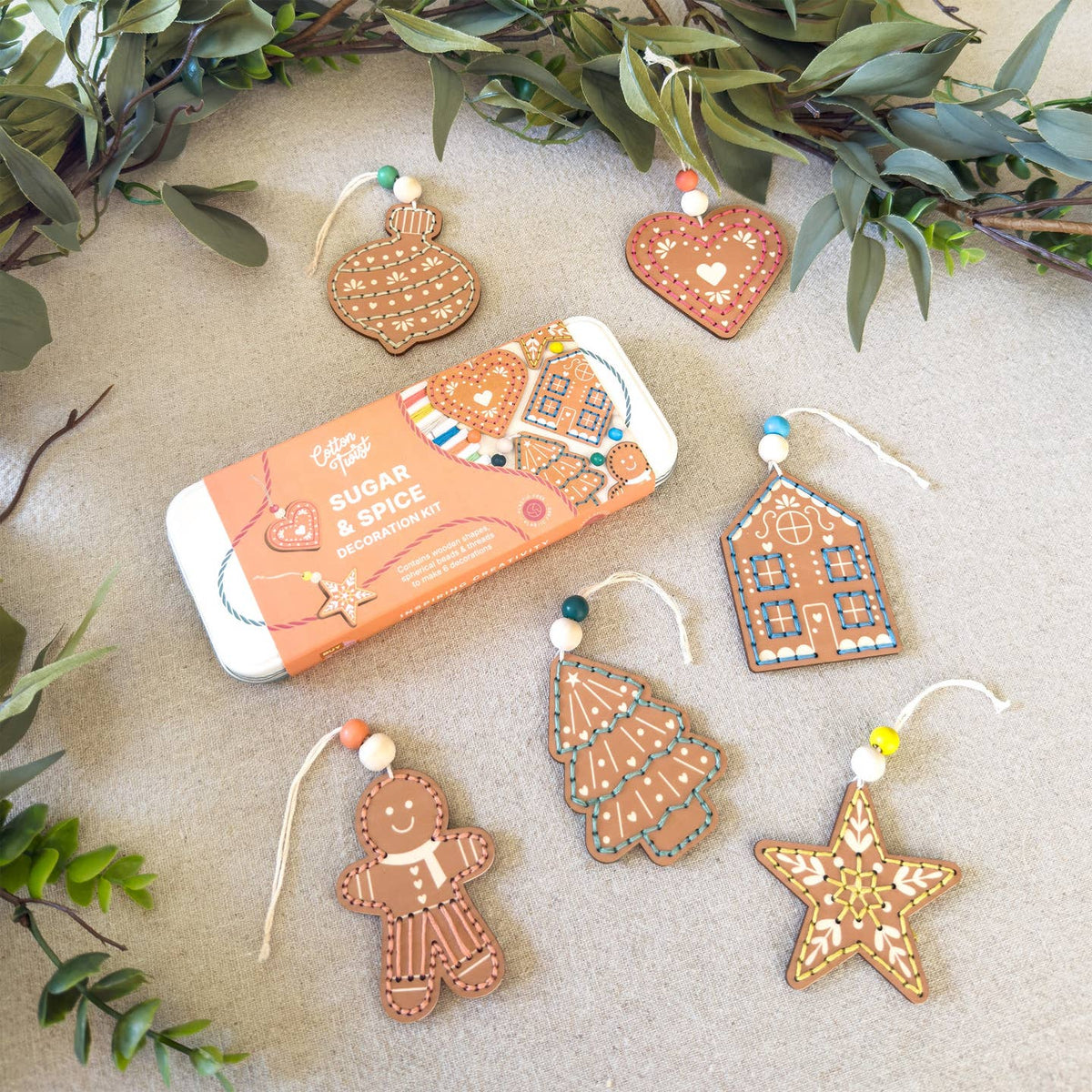 A tin of Sugar & Spice gingerbread ornament set arranged on a neutral surface with greenery. The set includes five decorated gingerbread ornaments shaped like a house, tree, star, gingerbread man, and Christmas tree, alongside the orange and white product tin.