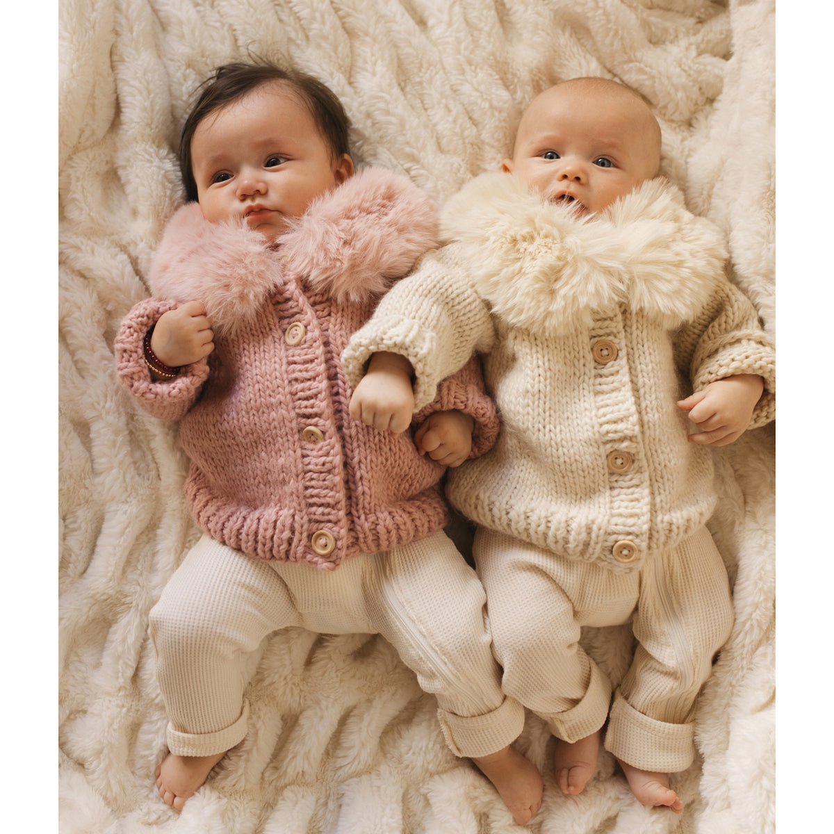 Two babies wearing knitted outfits with fluffy collars on a soft, textured surface.