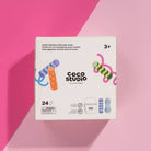A white box of Coco Studio Jumbo Rainbow Sidewalk Chalk by cocovillage, displayed on a pink and magenta background. The packaging features colorful illustrations of rainbow sidewalk chalk and text indicating it contains 24 pieces and is suitable for ages 3+.