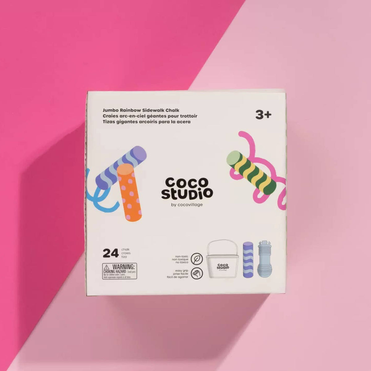 A white box of Coco Studio Jumbo Rainbow Sidewalk Chalk by cocovillage, displayed on a pink and magenta background. The packaging features colorful illustrations of rainbow sidewalk chalk and text indicating it contains 24 pieces and is suitable for ages 3+.