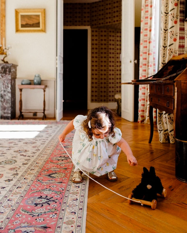 A young girl in a white floral dress crouches on a wooden floor, pulling a small black plush toy with a string in a vintage room with patterned rugs and wooden furniture.