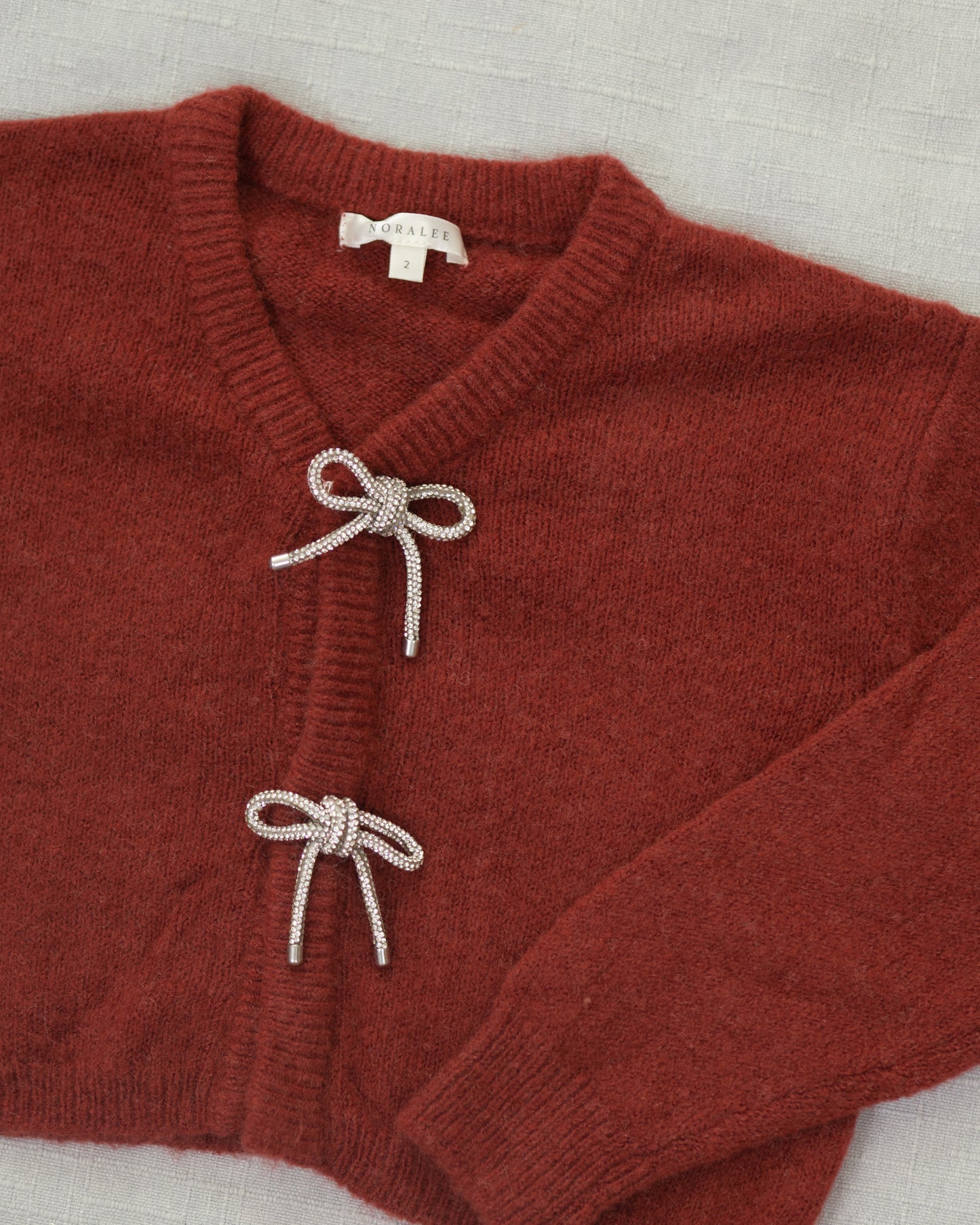 A close-up of a rust-red knit cardigan sweater with two decorative knotted bow details on the front, laid flat on a light textured fabric background. The brand name 'SARAH' is visible on the inner neck label.