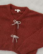 A close-up of a rust-red knit cardigan sweater with two decorative knotted bow details on the front, laid flat on a light textured fabric background. The brand name 'SARAH' is visible on the inner neck label.