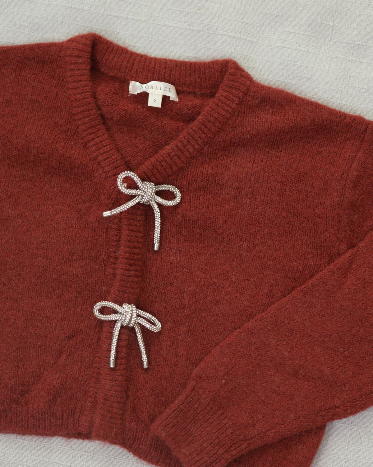 A close-up of a rust-red knit cardigan sweater with two decorative knotted bow details on the front, laid flat on a light textured fabric background. The brand name 'SARAH' is visible on the inner neck label.