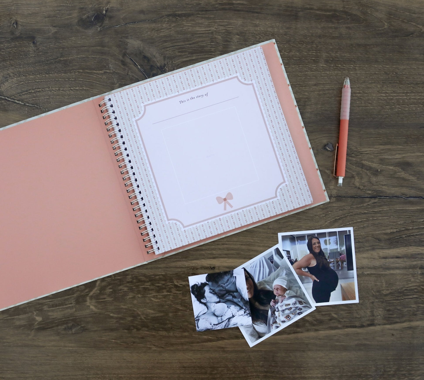 Open spiral-bound notebook with pink cover and white pages on a wooden surface, accompanied by a pen and two photos.