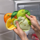 Hands holding silicone fruit-shaped food covers in front of a refrigerator.