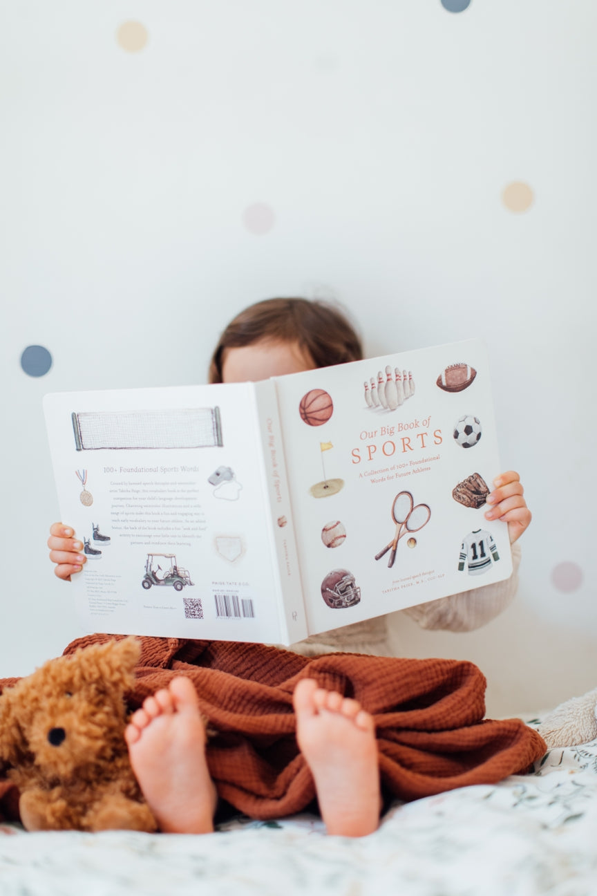 Child reading a book titled 'Sports' with a teddy bear on a bed.