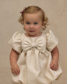 A toddler girl with curly light brown hair wearing a cream-colored textured dress with a large bow at the chest and puffed short sleeves, standing against a neutral beige background.