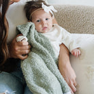 Baby wrapped in a green blanket with a white headband, held by an adult on a beige couch.