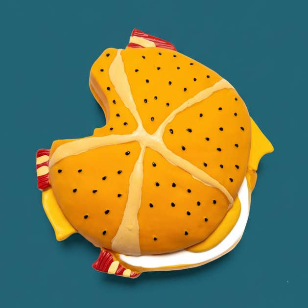 A novelty cake shaped like a hamburger, with a golden-brown bun decorated with black sesame seeds, cream-colored frosting lines, yellow cheese layers, and red-and-white striped buns, presented on a solid teal background.
