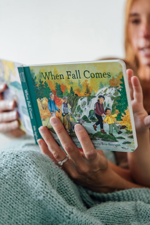A toddler holding an open board book titled When Fall Comes, looking down at the colorful illustrated pages showing autumn trees and an outdoor nature scene.